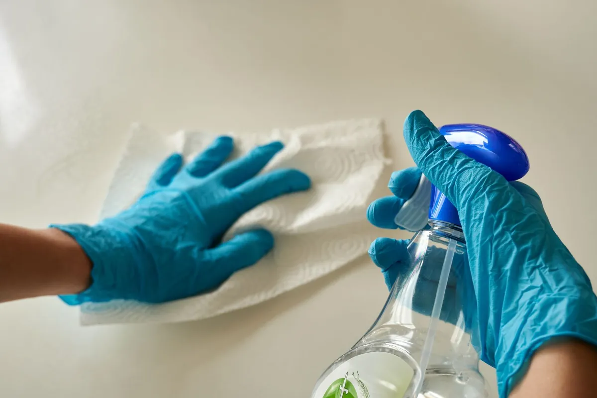 Blue gloved hands wiping a white surface while holding a spray bottle