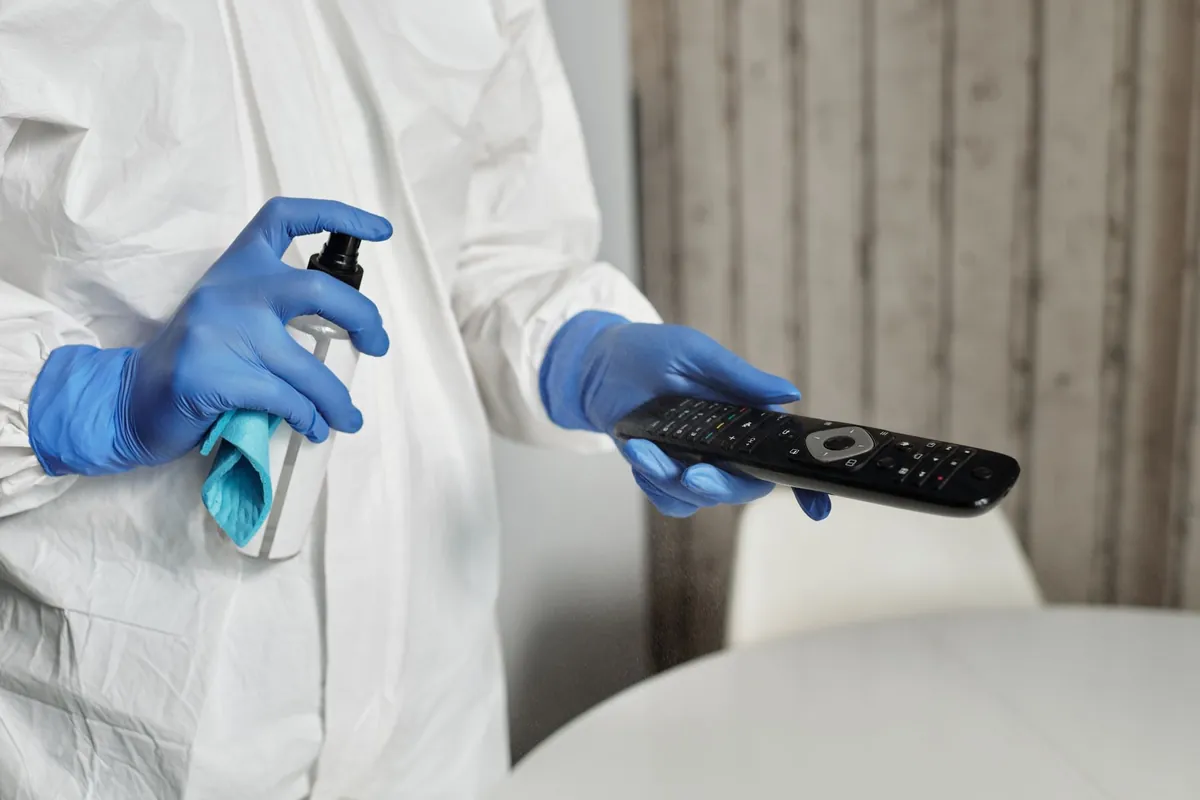Person in a white protective suit with blue gloves spraying and wiping a remote control (torso only)